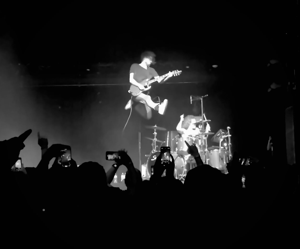 A guitarist mid-jump above the drum kit, seen from a crowd holding up phones